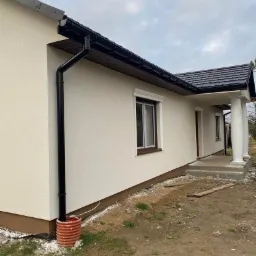 Wykończona elewacja domu jednorodzinnego z kremowym tynkiem, czarną rynną i brązowymi ramami okiennymi, widoczna pomarańczowa studzienka kanalizacyjna przy ścianie.