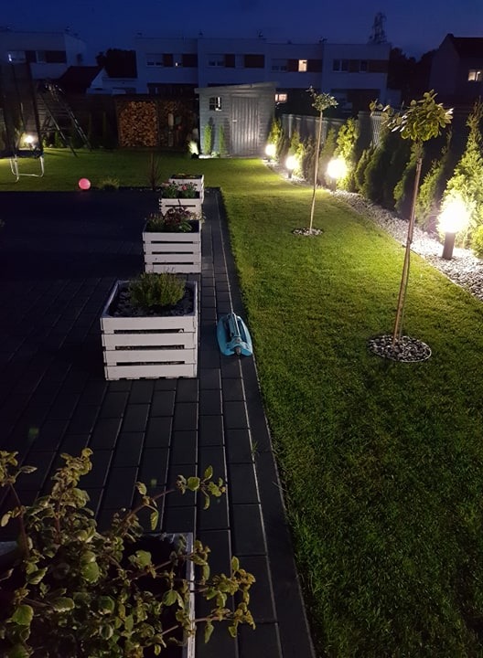 Oświetlony ogród nocą z trawnikiem, białymi donicami z kwiatami, drzewkami ozdobnymi i automatycznym zraszaczem leżącym na kostce brukowej.