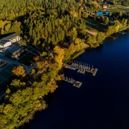 Widok z lotu ptaka na ośrodek wypoczynkowy nad jeziorem z pomostem dla żaglówek, kortami tenisowymi i budynkiem hotelowym otoczonym jesiennym lasem.
