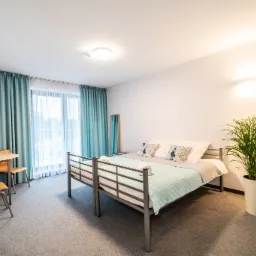 Pokój z łóżkiem piętrowym, turkusową narzutą i poduszkami z motywem pawich piór, stolikiem z owocami, zasłonami w odcieniach turkusu oraz rośliną doniczkową w białej osłonce.