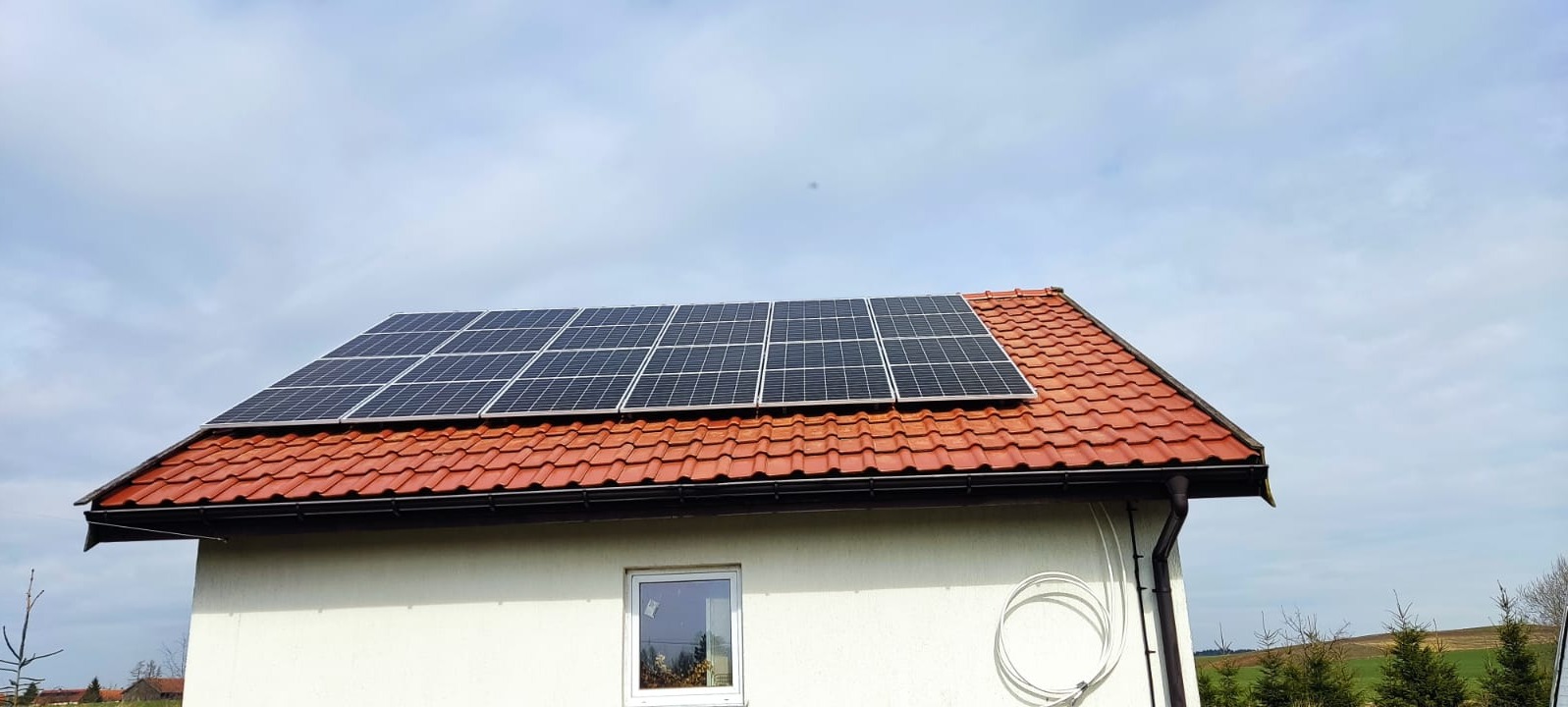 Panel słoneczny na dachu domu jednorodzinnego z czerwoną dachówką, widok z dołu na tle pochmurnego nieba.