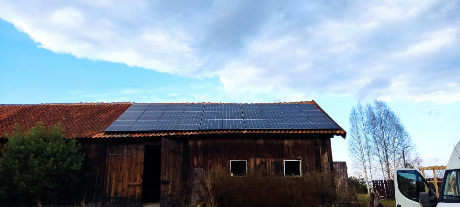 Panele słoneczne na dachu starej, drewnianej stodoły z czerwoną dachówką, na tle błękitnego nieba z chmurami. Częściowo otwarte wrota stodoły.