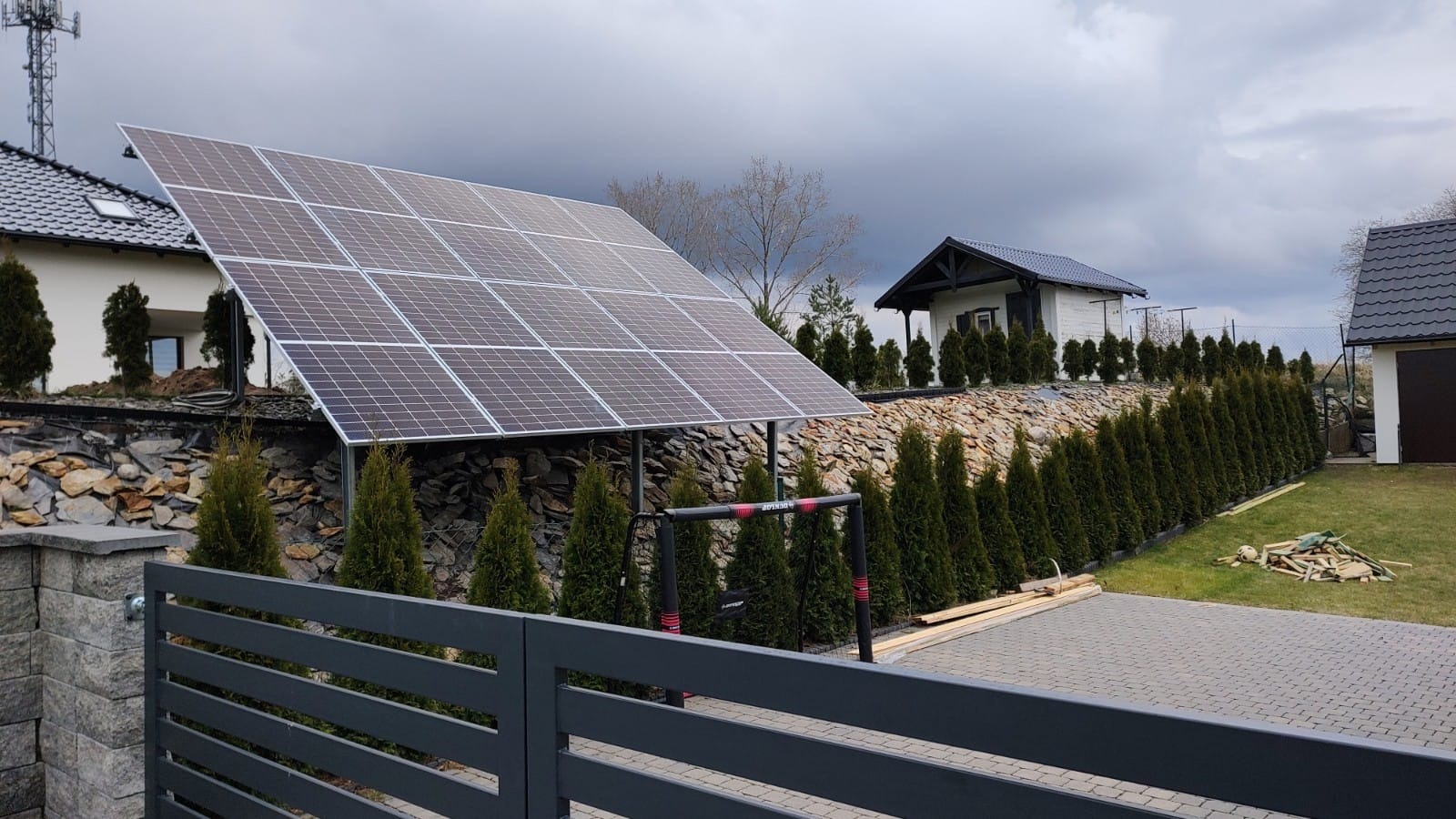Instalacja paneli słonecznych na metalowej konstrukcji, widok zza szarego ogrodzenia, w tle dom i tuje na kamiennym murku, pochmurne niebo.