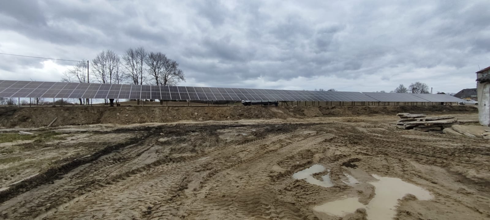 Długa instalacja fotowoltaiczna zamontowana na konstrukcji nad ziemią, widoczne ślady opon w błotnistym terenie i sterta płyt betonowych