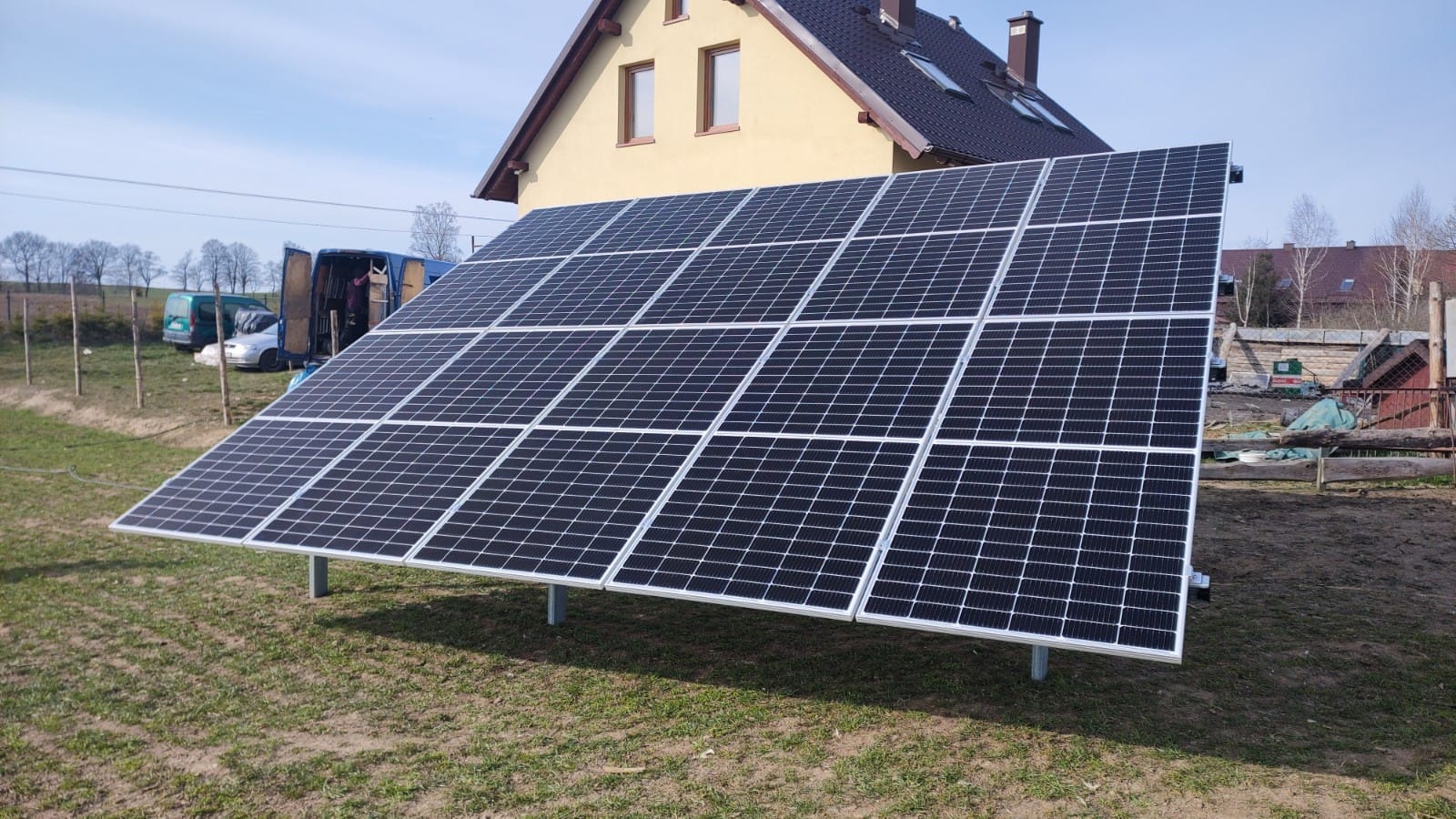 Panel fotowoltaiczny zainstalowany na gruncie, widoczny dom jednorodzinny w tle, dwa busy instalacyjne zaparkowane obok ogrodzenia.