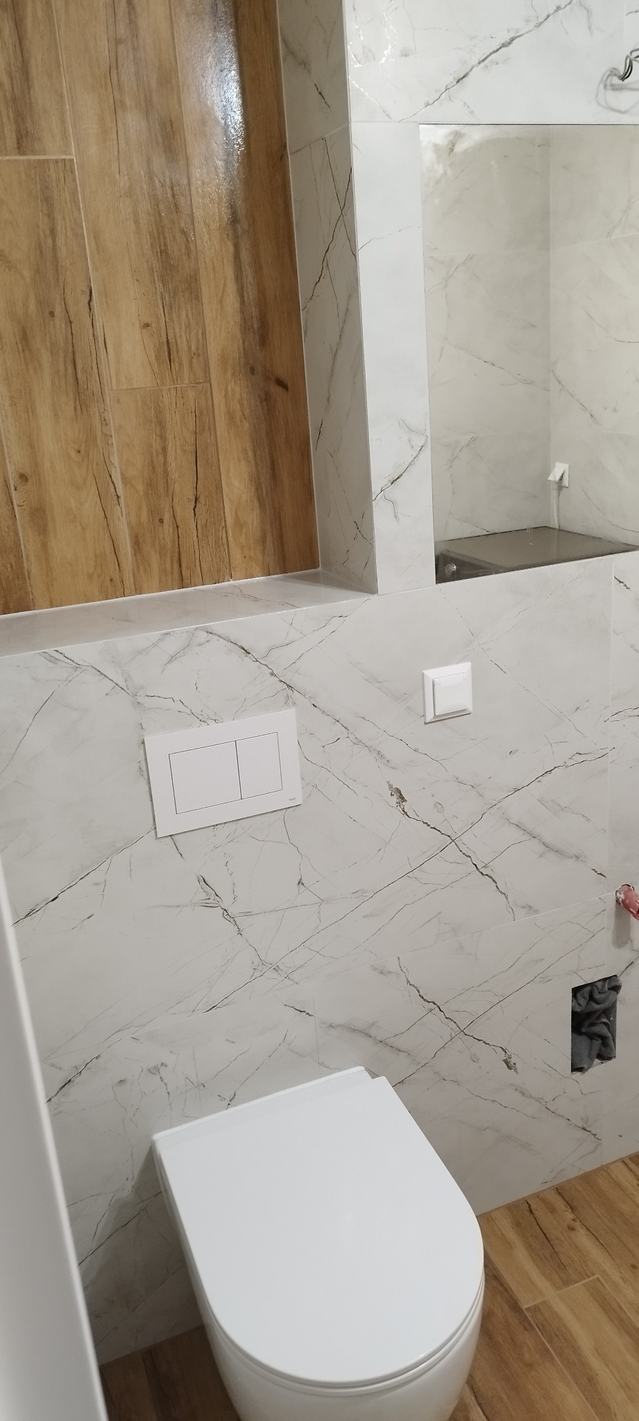 Nowoczesna łazienka z białymi płytkami imitującymi marmur, drewnopodobną podłogą, wiszącą miską WC i ukrytym systemem spłukiwania.