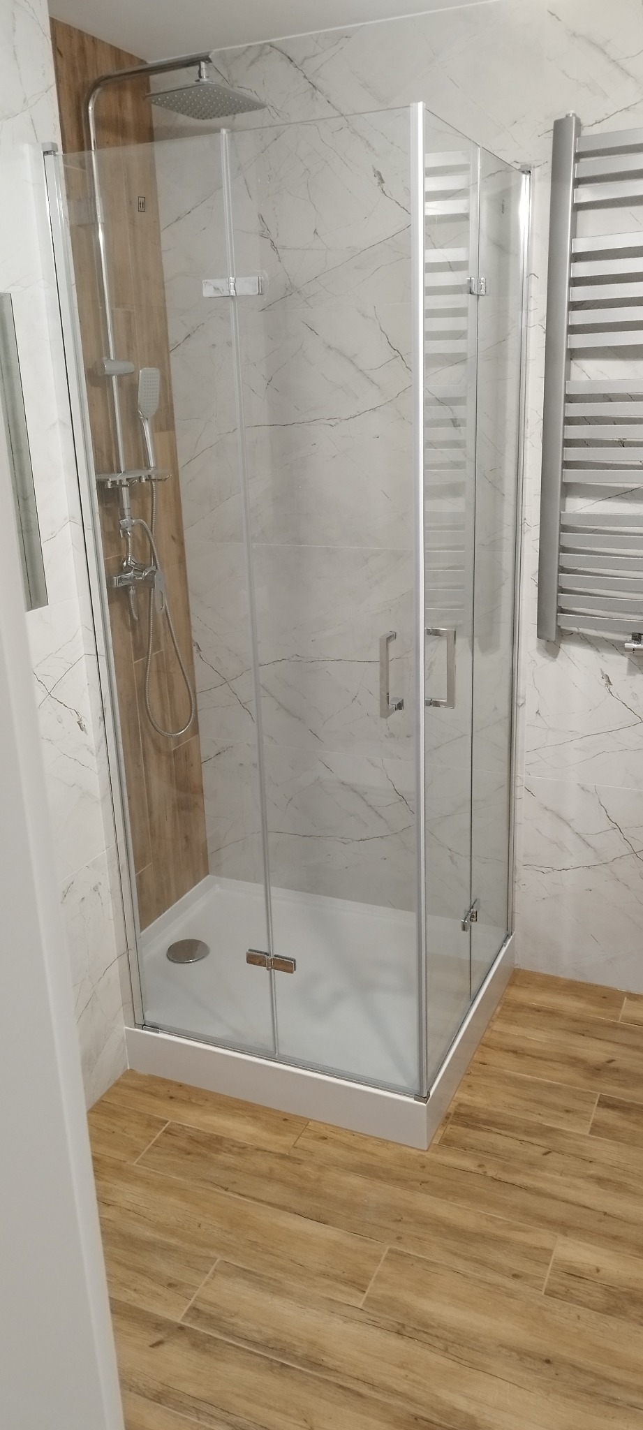Nowoczesna kabina prysznicowa z deszczownicą i panelem prysznicowym, wykończona płytkami imitującymi marmur i drewno, z grzejnikiem drabinkowym w tle.