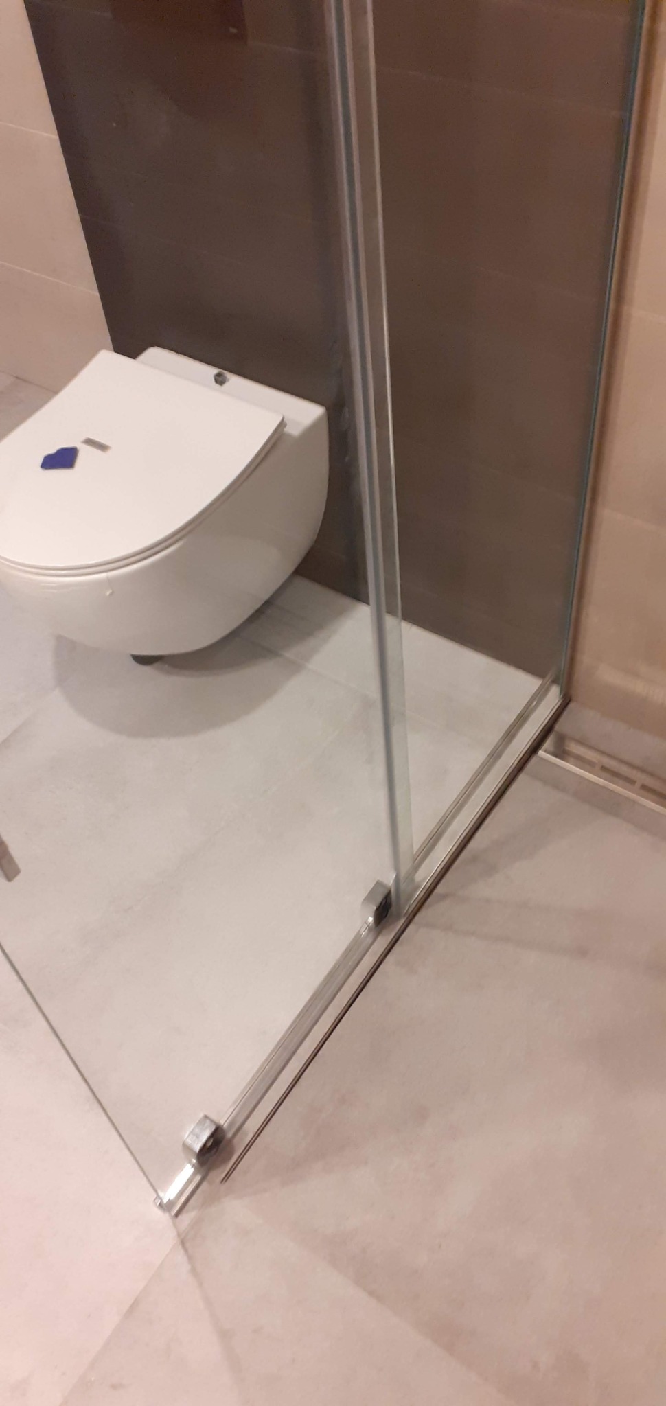 Nowoczesna, podwieszana toaleta z deską wolnoopadającą w minimalistycznej łazience za szklaną kabiną prysznicową, na jasnych płytkach podłogowych.