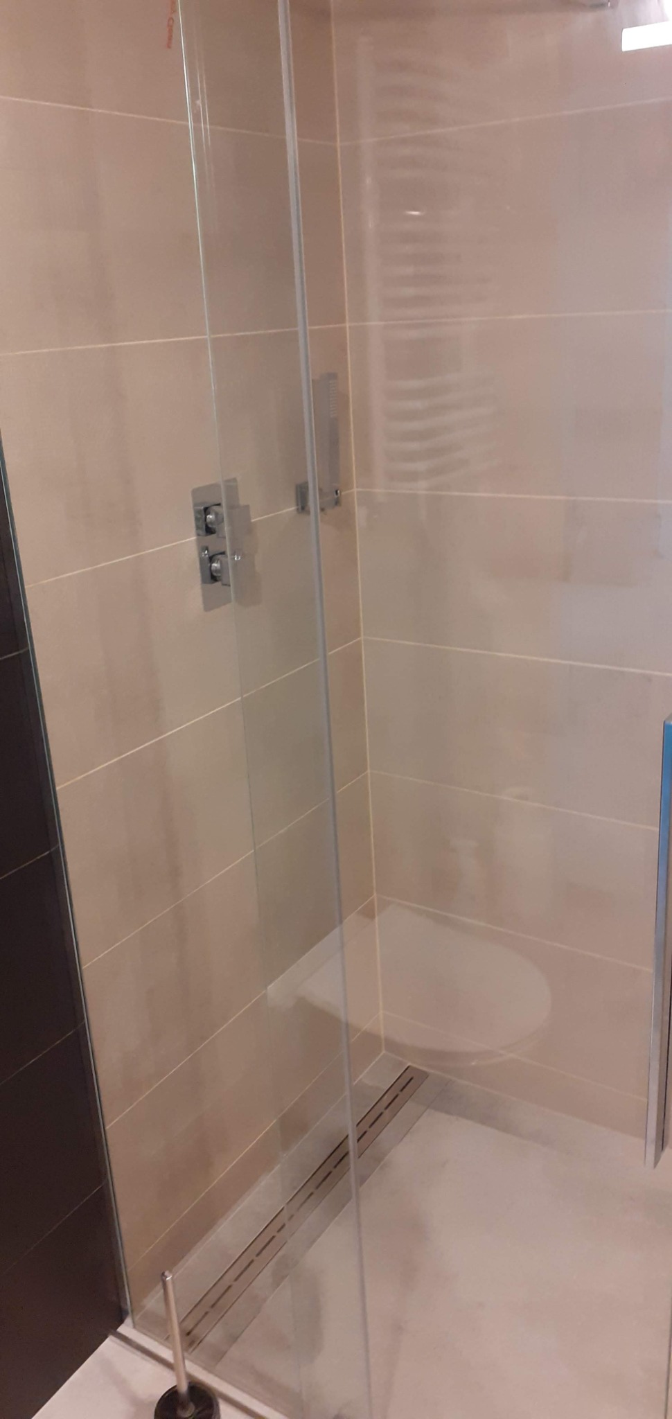 Szklana kabina prysznicowa z zamontowanym siedziskiem i odpływem liniowym, wykończona beżowymi płytkami ceramicznymi z chromowaną armaturą.