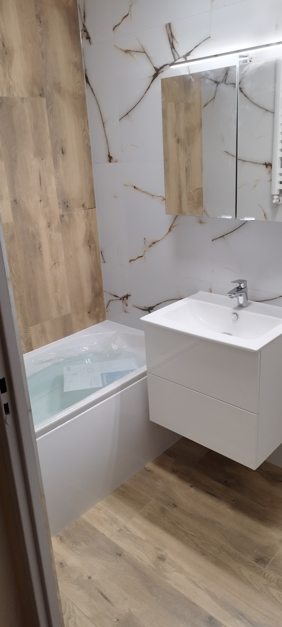 Nowoczesna łazienka z wanną w folii ochronnej, białą szafką pod umywalką i płytkami imitującymi drewno na podłodze i ścianie, uzupełniona białymi płytkami z marmurkowym wzorem.