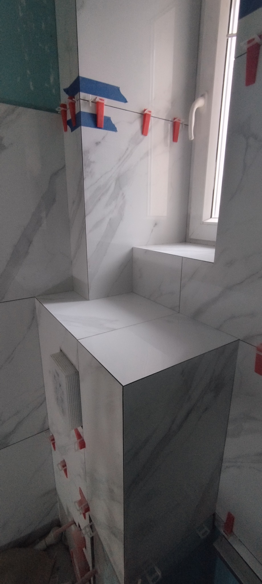 Świeżo ułożone, białe płytki imitujące marmur z szarymi żyłami na ścianie i obudowie wnęki okiennej w trakcie prac, widoczne czerwone klipsy i niebieska taśma poziomująca.