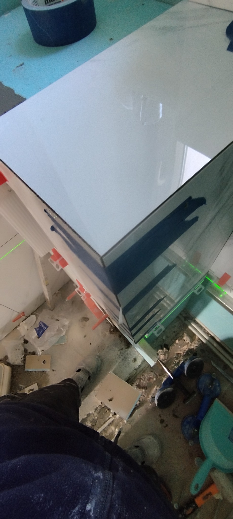 Układanie białych płytek w rogu łazienki, widoczne poziomice laserowe i narzędzia glazurnicze, w tle nieporządek na podłodze.