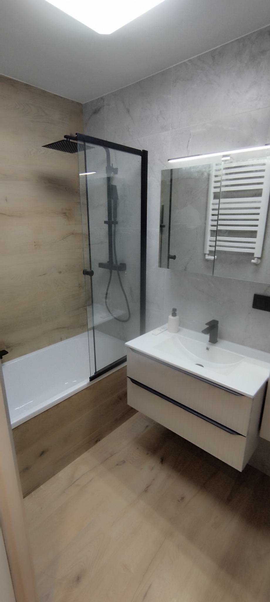 Nowoczesna łazienka z kabiną prysznicową typu walk-in z czarnymi profilami, białą umywalką nablatową z czarną baterią oraz grzejnikiem drabinkowym.