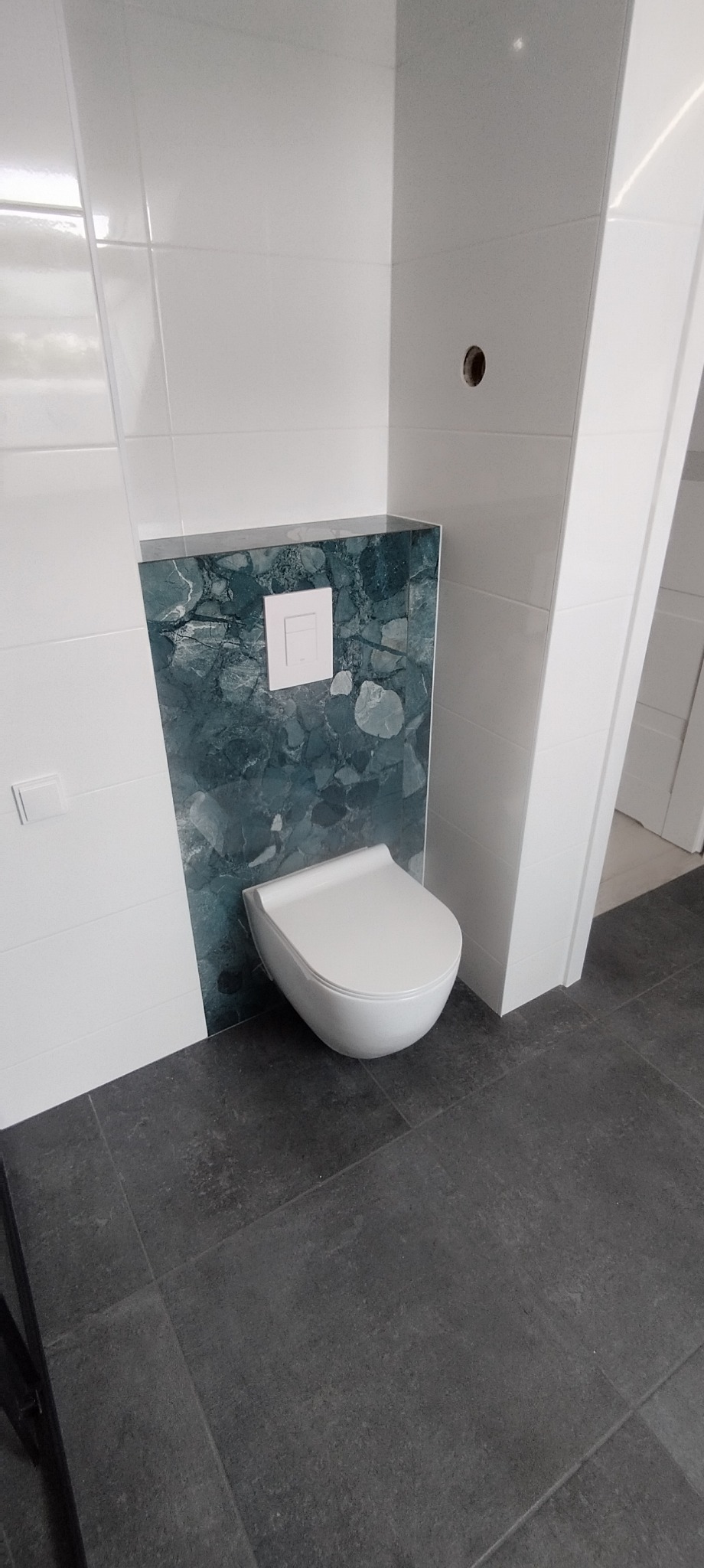 Nowoczesna toaleta podwieszana z marmurową obudową spłuczki i białymi płytkami na ścianach, szare płytki podłogowe, minimalistyczny design.