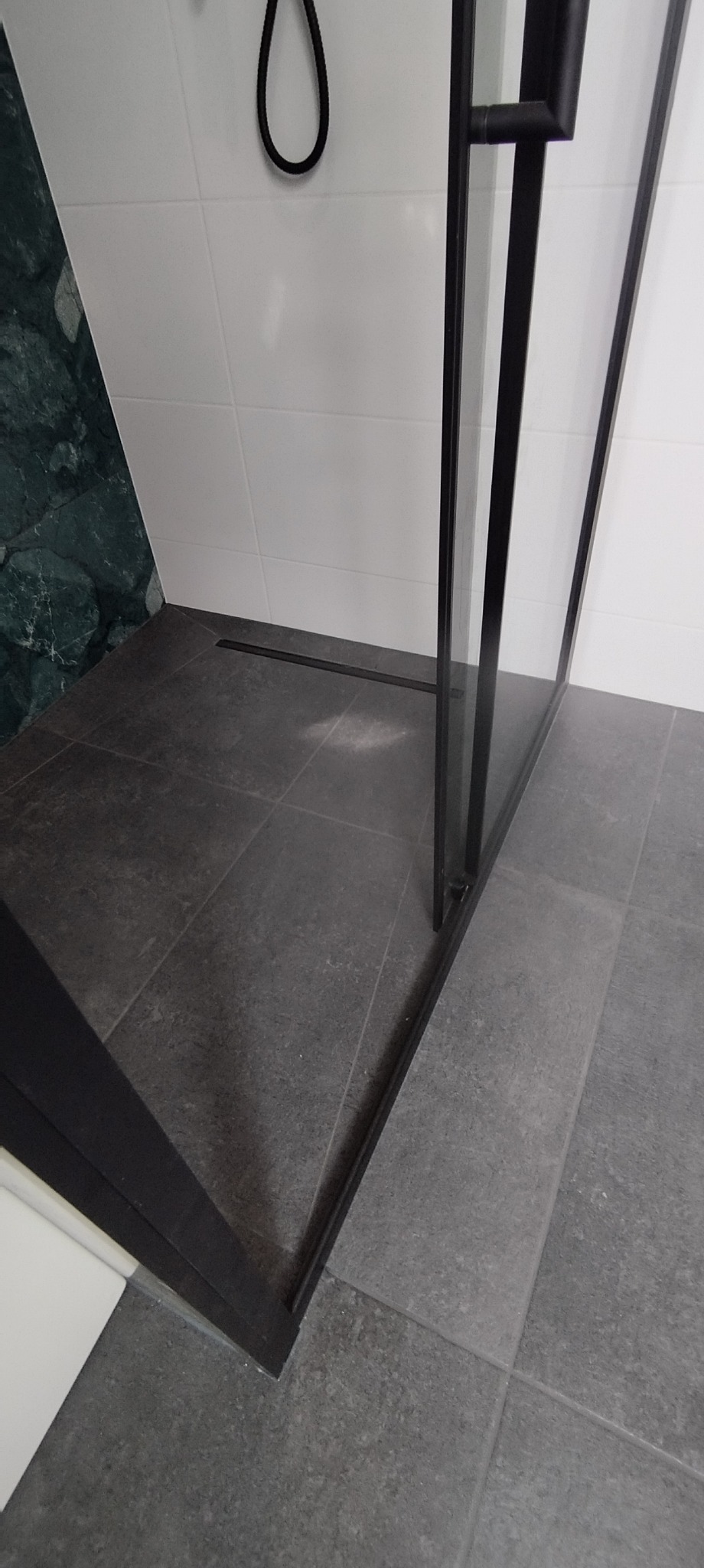 Nowoczesna kabina prysznicowa z czarnymi profilami, szare płytki podłogowe i białe płytki ścienne z elementem zielonego marmuru. Minimalistyczny design i eleganckie wykończenie.