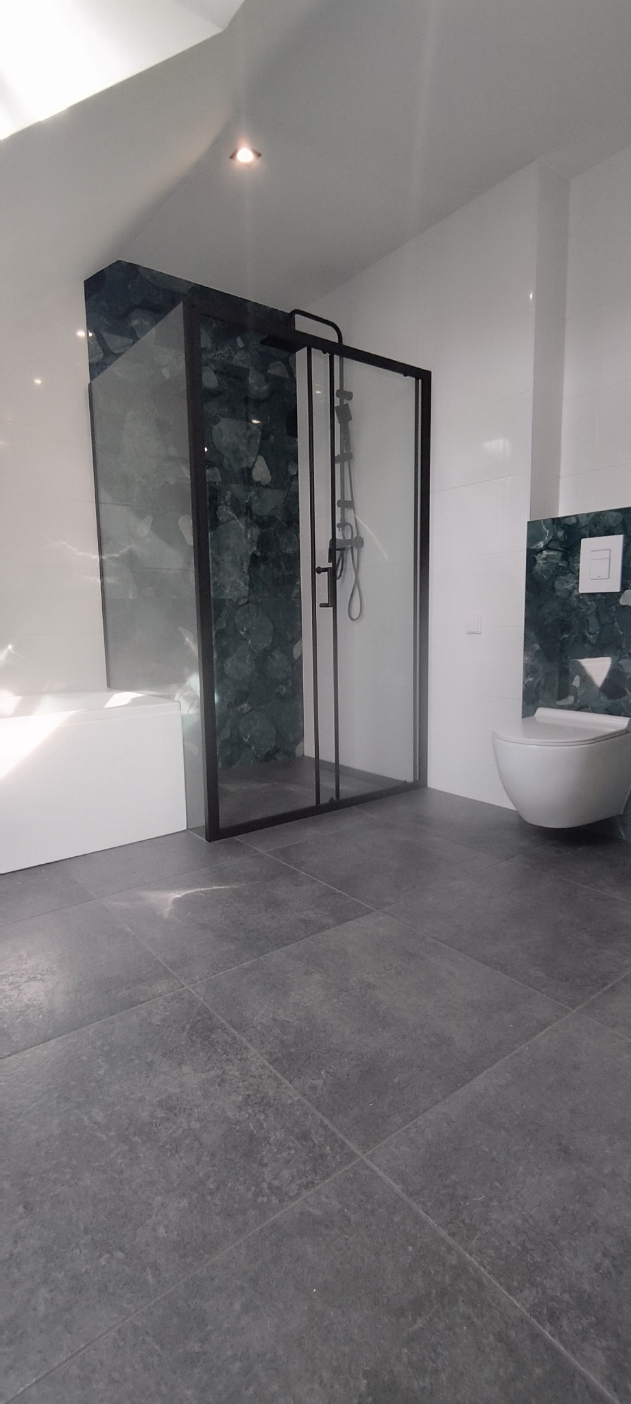 Nowoczesna łazienka z kabiną prysznicową typu walk-in z czarnymi ramami, szare płytki podłogowe i akcenty z zielonego marmuru na ścianach.