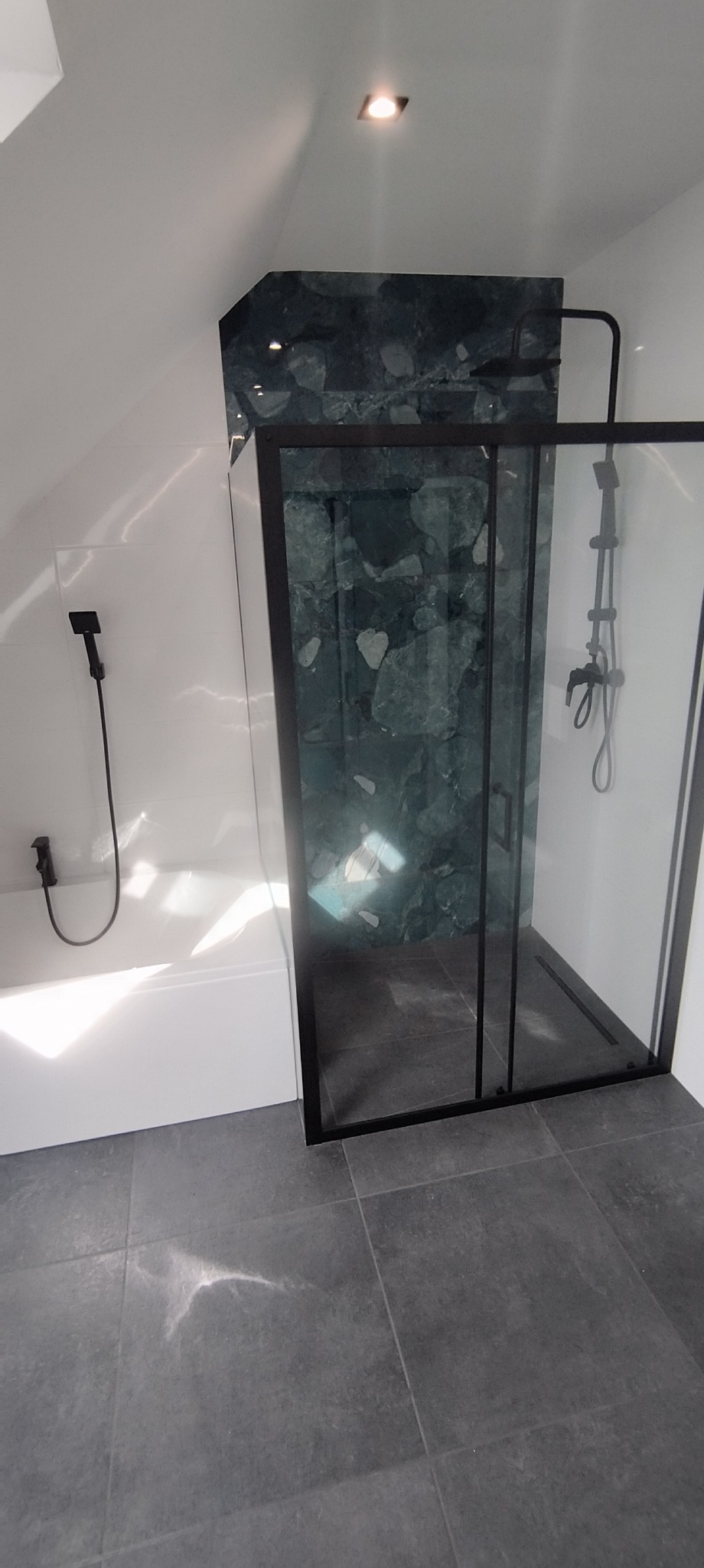 Nowoczesna łazienka na poddaszu z kabiną prysznicową typu walk-in, wykończona płytkami imitującymi kamień, czarna armatura i szara podłoga.