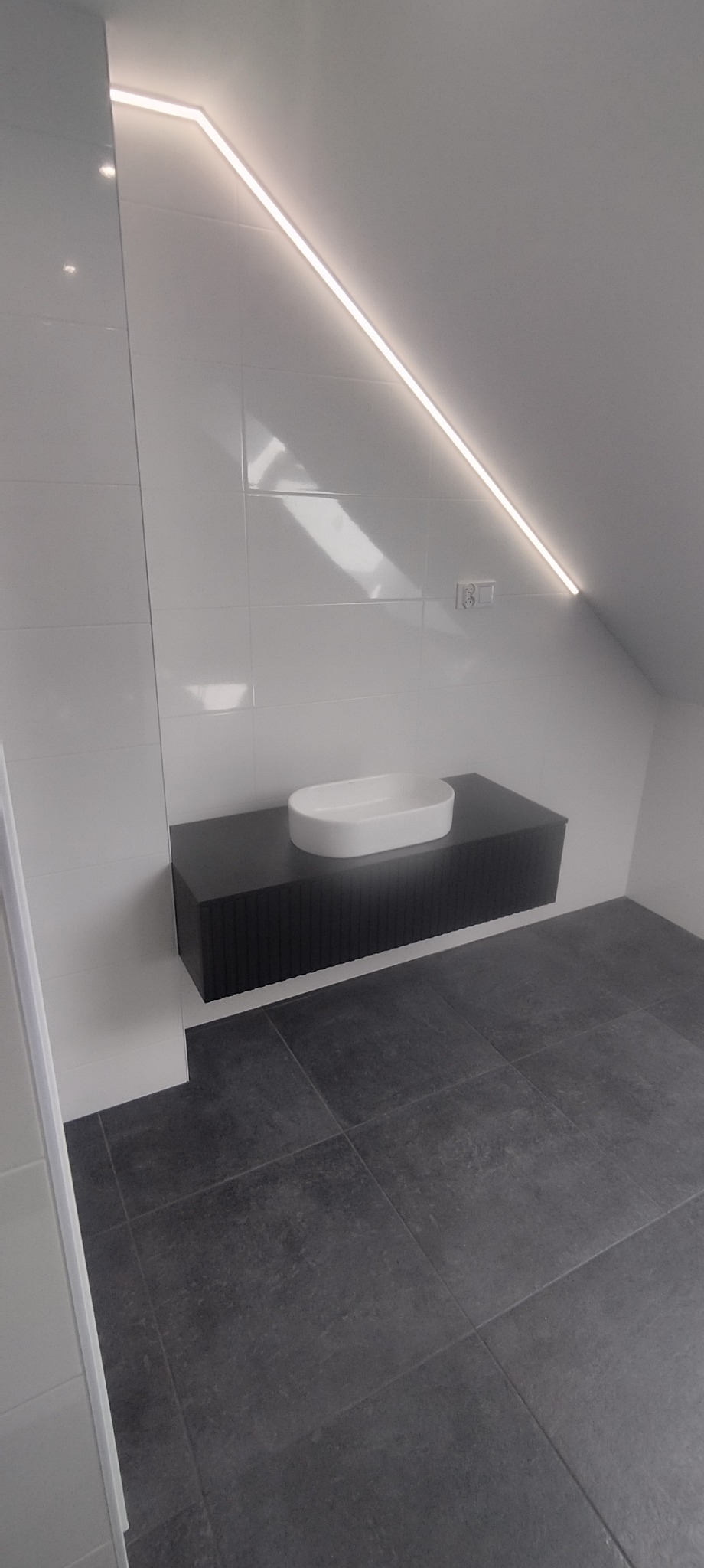 Nowoczesna łazienka na poddaszu: białe płytki, czarna szafka z umywalką, szara podłoga i oświetlenie LED wzdłuż skosu. Minimalistyczny design.