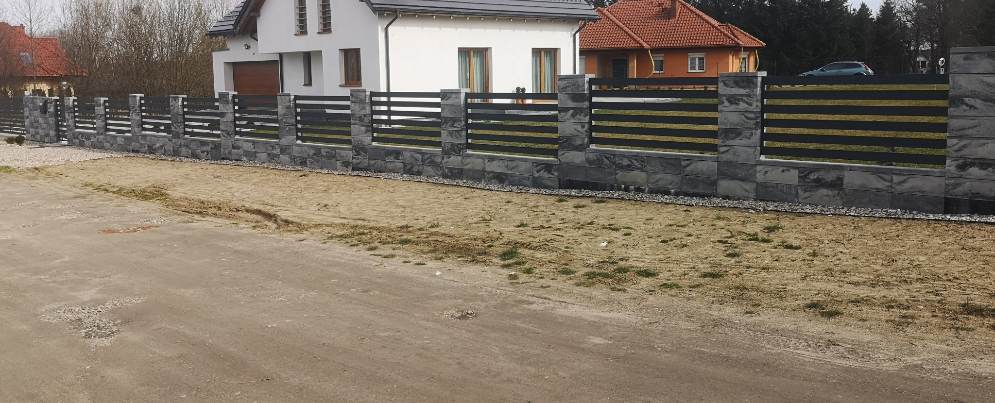 Nowoczesne ogrodzenie z betonowych bloczków i poziomych, czarnych przęseł metalowych, z widocznymi domami w tle.