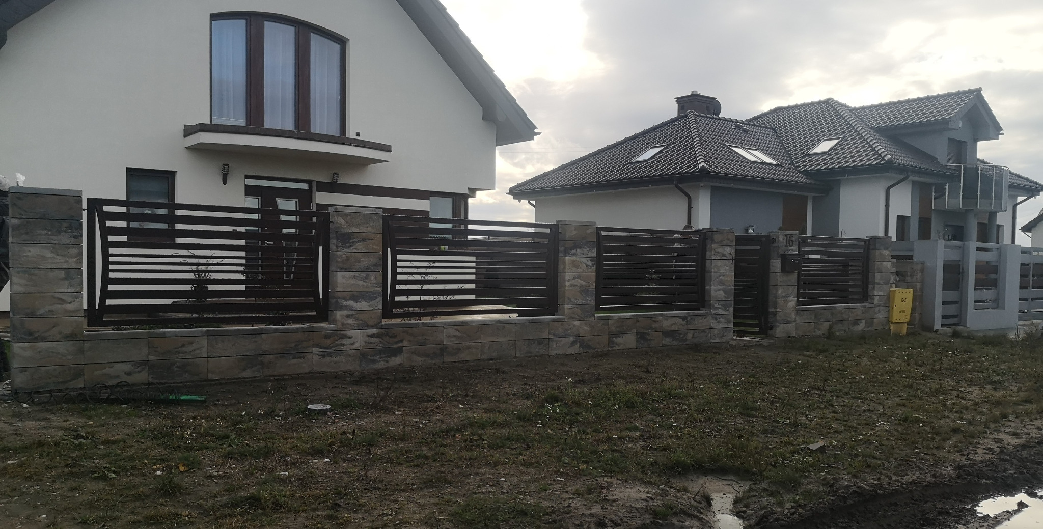 Nowoczesne ogrodzenie posesji z poziomych, ciemnych paneli metalowych, osadzone na niskiej podmurówce i słupkach z szarej kostki imitującej kamień. W tle widoczne dwa domy jednorodzinne.