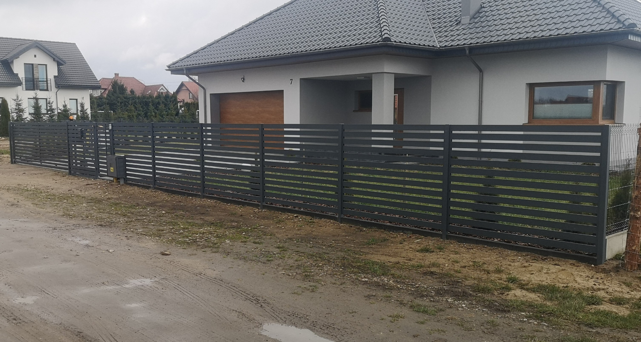 Nowoczesne, poziome, szare ogrodzenie metalowe z szerokimi panelami, z przesuwną bramą, na tle domu z szarą dachówką i brązową bramą garażową, widok z zewnątrz na mokrą, gruntową drogę.