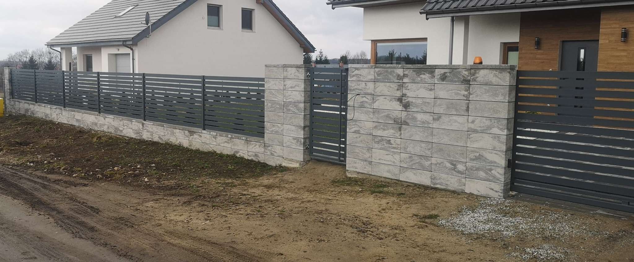 Nowoczesne ogrodzenie posesji z szarych paneli poziomych i słupków z betonowych bloczków imitujących kamień, z bramą wjazdową przesuwną, na tle dwóch budynków mieszkalnych.
