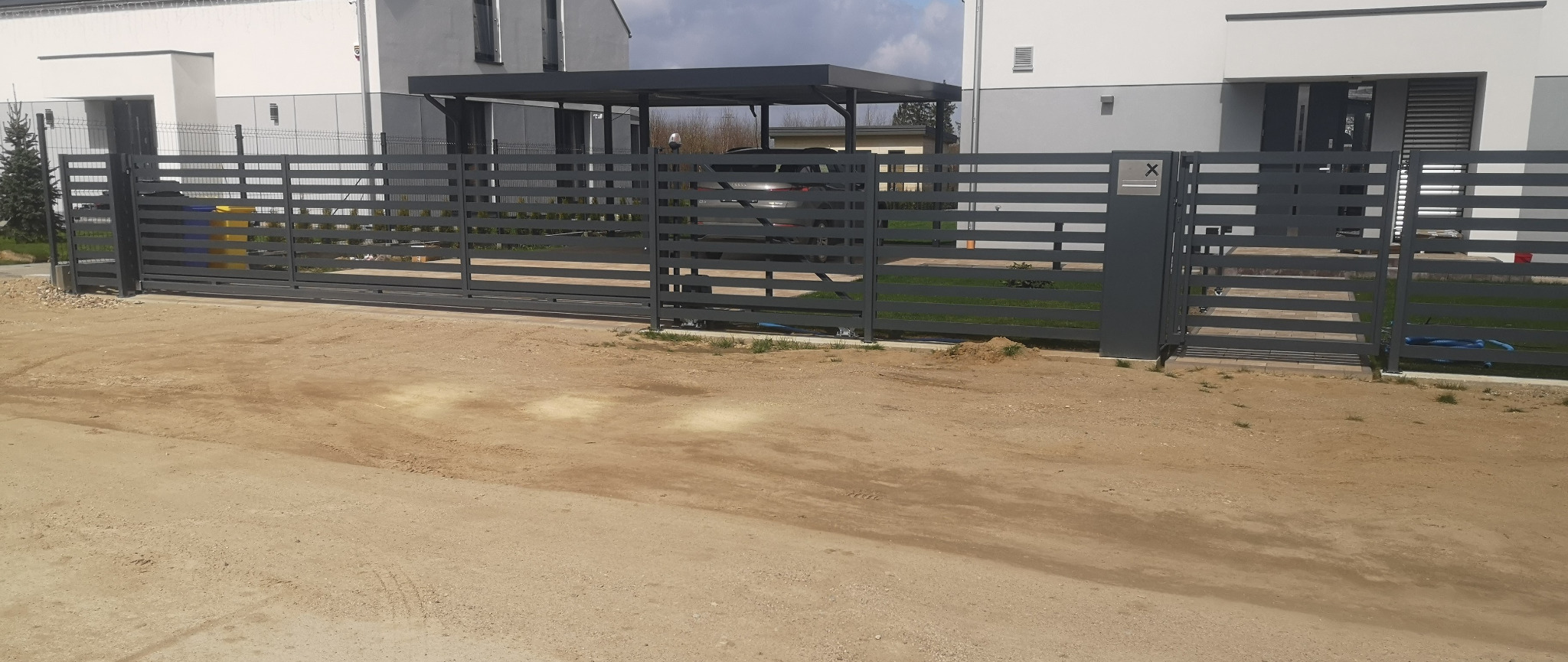 Nowoczesna, przesuwna brama wjazdowa z poziomych profili metalowych w kolorze grafitowym, z furtką po prawej stronie i panelem z numerem domu, na tle nowego domu z carportem i niezagospodarowanego...