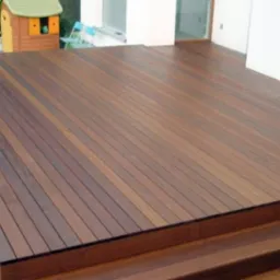 Ciemnobrązowy taras drewniany z widocznymi schodami, przylegający do białej ściany budynku, z małym domkiem dla dzieci w tle.