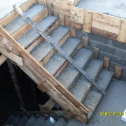 Betonowe schody w trakcie budowy, z drewnianym szalunkiem, widoczne bloczki fundamentowe i data na zdjęciu.