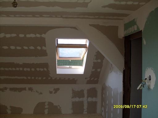 Poddasze w trakcie remontu: ściany przygotowane do malowania, widoczne paski szpachli, okno dachowe z roletą, surowy stan wykończenia.