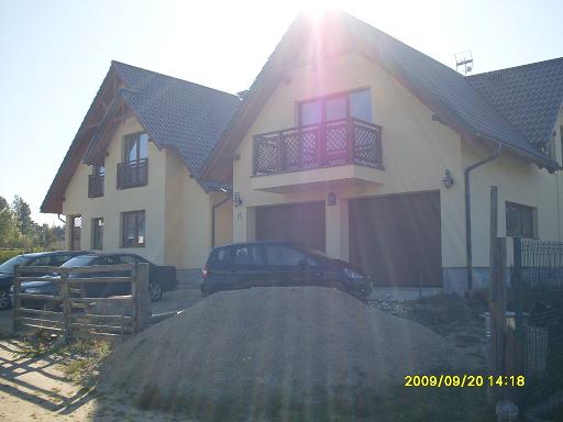 Dwa budynki mieszkalne z garażami, widoczne samochody i sterta piasku na pierwszym planie, jasna elewacja i ciemna dachówka, słoneczny dzień.