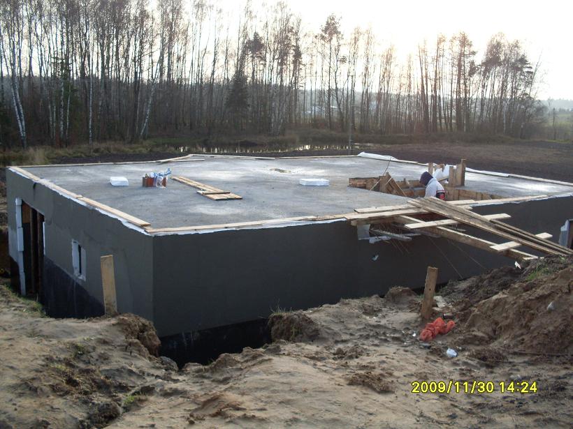 Budowa domu w stanie surowym zamkniętym: fundament, ściany z ociepleniem i strop, widoczne drewniane szalunki i materiały budowlane na placu budowy, w tle drzewa.