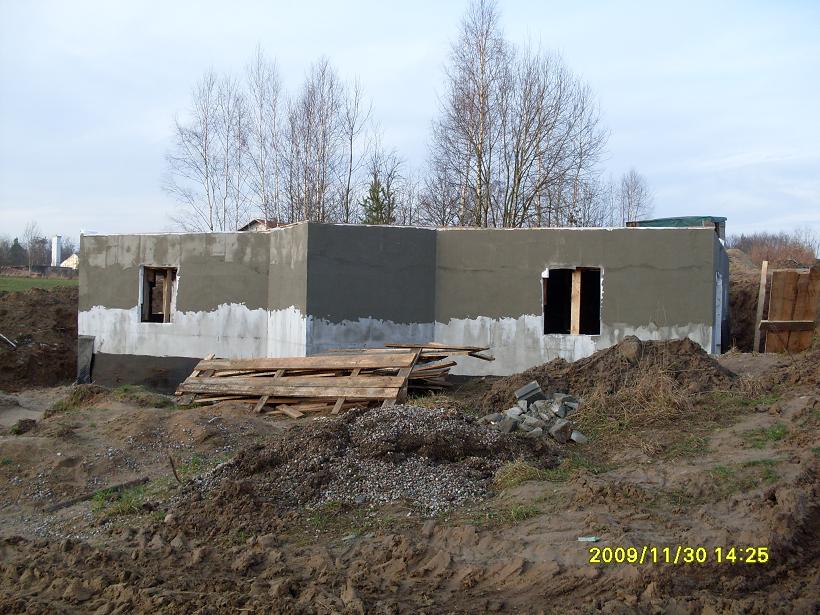 Surowy stan ścian domu murowanego z otworami okiennymi, widoczne palety i gruz na placu budowy.