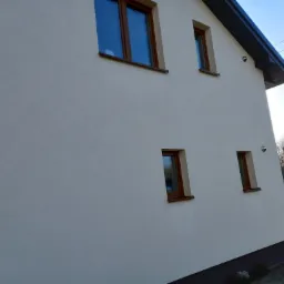 Jasna elewacja domu jednorodzinnego z drewnianą stolarką okienną, widok z boku, w tle zaparkowany samochód i linia drzew.