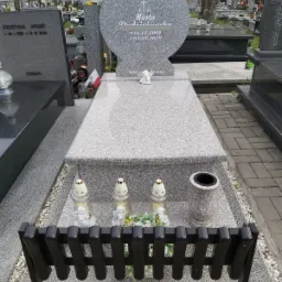 Granitowy nagrobek w kształcie serca z inskrypcją 'Ś.P. Marta Podniesińska 16.12.2002 +03.05.2019 Dzieli na tylko czas', ozdobiony figurką anioła, lampionami i kwiatami, widok zza czarnej ławeczki.