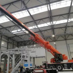 Pomarańczowy dźwig samochodowy marki Liebherr z wysięgnikiem teleskopowym, z napisem WOJRES, podnosi element konstrukcyjny wewnątrz hali przemysłowej z widoczną stalową konstrukcją dachu.