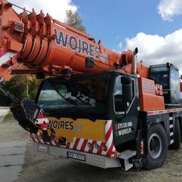 WOJ-RES, Adam Wojnar - Pomarańczowy dźwig samochodowy firmy Wojres zaparkowany na piaszczystym terenie z widokiem na jezioro i zalesione wzgórza w tle, widoczne numery kontaktowe i adres strony internetowej na kabinie.