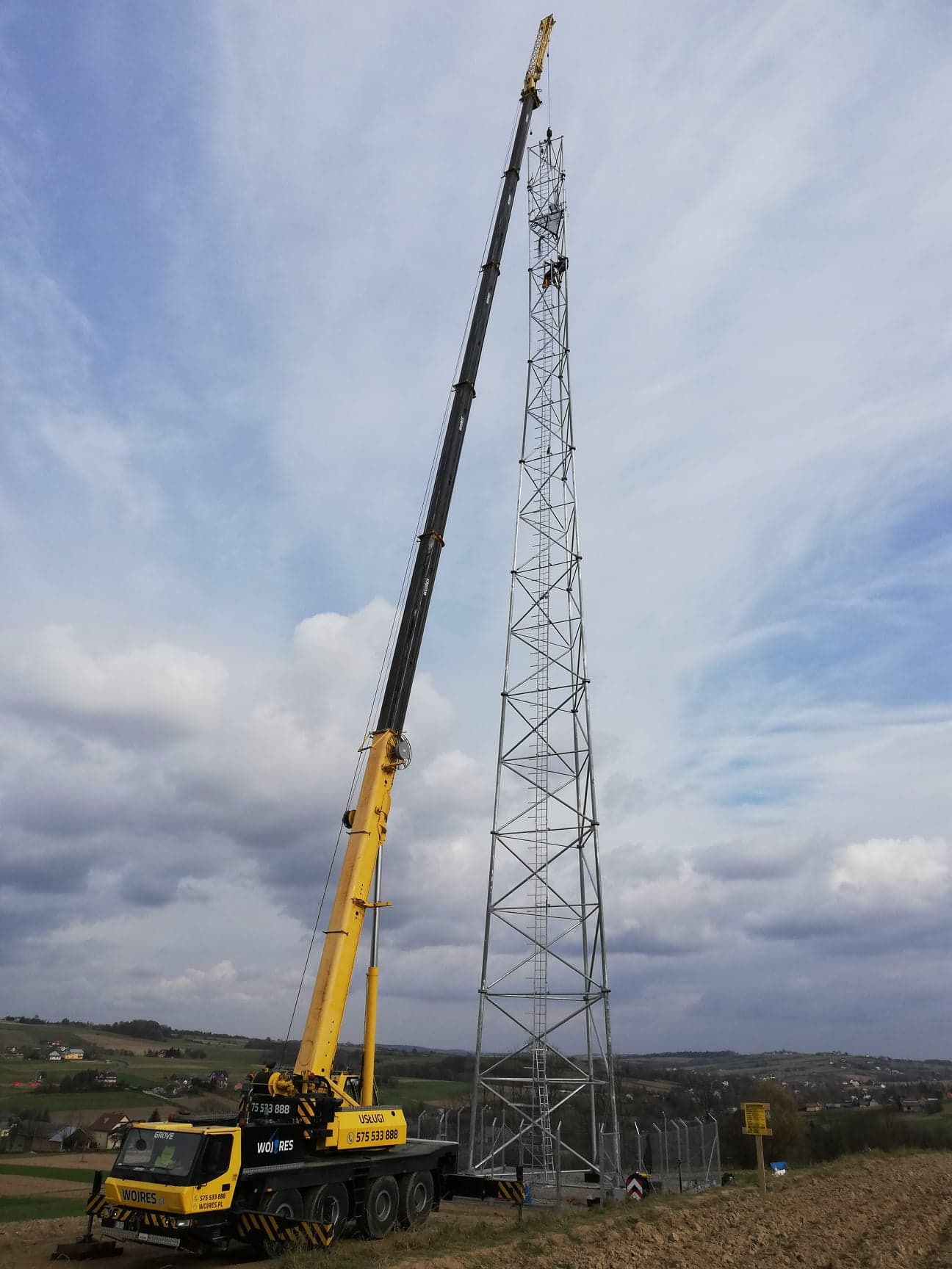 Żółty dźwig teleskopowy GROVE podnosi element stalowej konstrukcji wieży telekomunikacyjnej, widoczni pracownicy na wysokości, w tle pagórkowaty krajobraz pod pochmurnym niebem.
