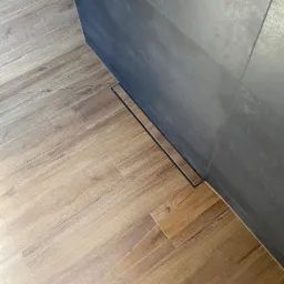 Ułożone pod kątem płytki podłogowe imitujące drewno w jasnych odcieniach brązu, stykające się z ciemnoszarą ścianą wyłożoną płytkami i liniowym odpływem podłogowym.