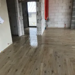 Świeżo ułożona, błyszcząca podłoga z paneli w jodełkę w surowym wnętrzu z białych bloczków betonowych i widocznymi kablami elektrycznymi.