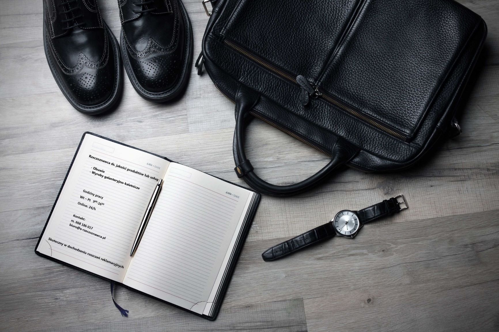 Eleganckie czarne buty, skórzana torba, zegarek i otwarty notatnik z zapisanymi danymi kontaktowymi rzeczoznawcy na drewnianym tle.