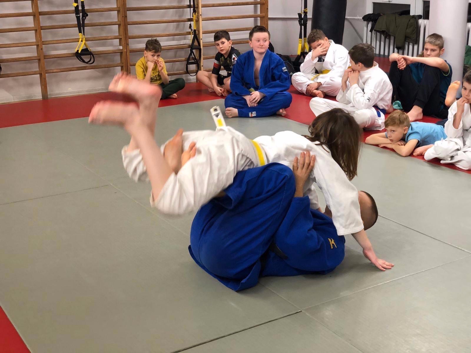 Dzieci ćwiczą rzut podczas treningu judo, grupa obserwuje z boku, sala z drabinkami gimnastycznymi w tle.
