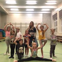 Grupa dzieci i instruktorka w sali gimnastycznej pozują do zdjęcia, wykonując figury gimnastyczne. Dziewczynka na pierwszym planie wykonuje szpagat.