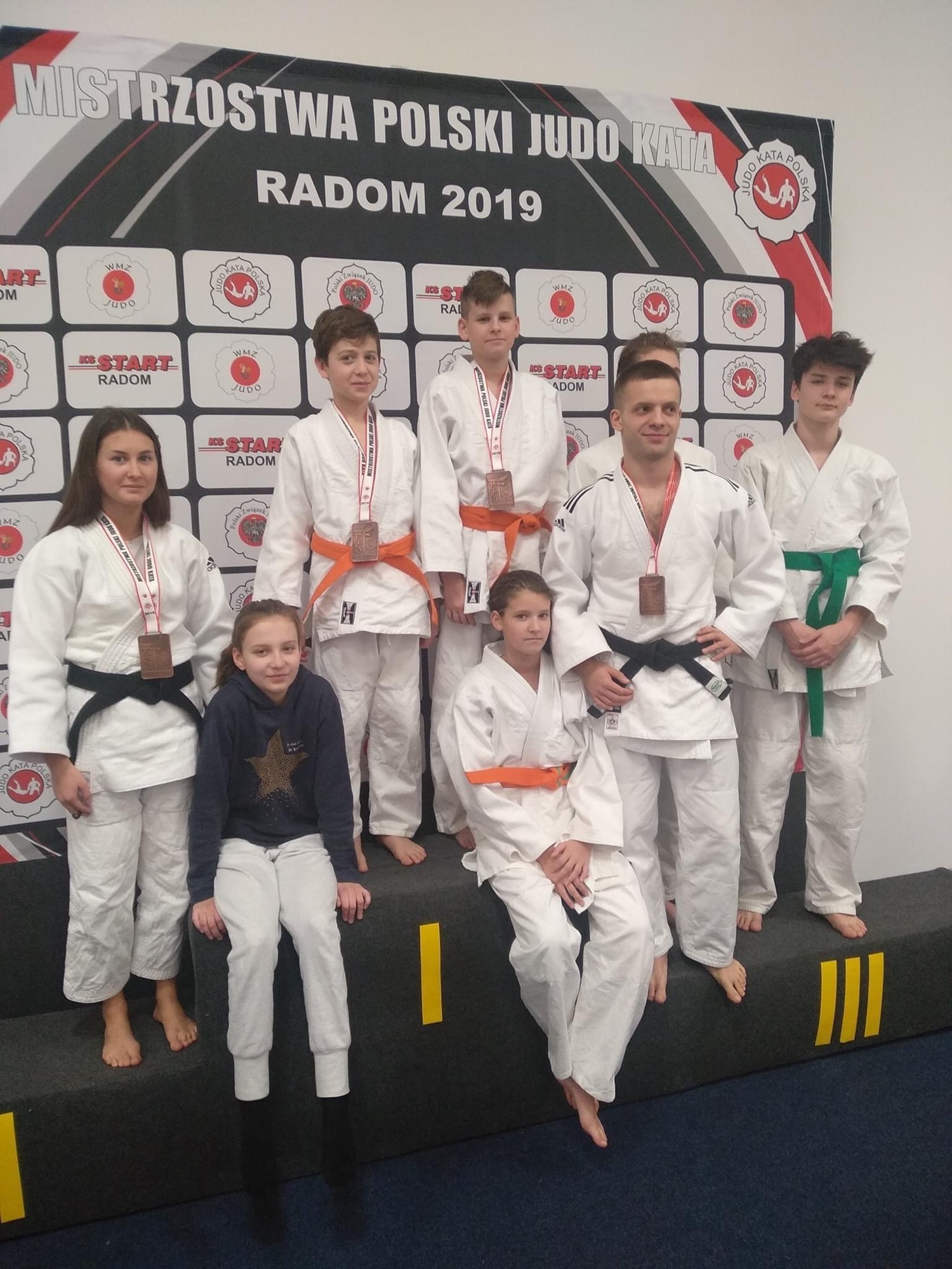 Grupa młodych judoków i instruktor na podium po Mistrzostwach Polski Judo Kata w Radomiu 2019, z różnymi pasami i brązowymi medalami.