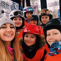 Grupa dzieci i dorosła kobieta w strojach narciarskich, uśmiechnięte twarze, widok z wnętrza kolejki linowej na ośnieżony stok.