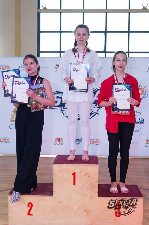 Trzy młode kobiety na podium z dyplomami i medalami, po zawodach tanecznych lub gimnastycznych, na tle baneru reklamowego.