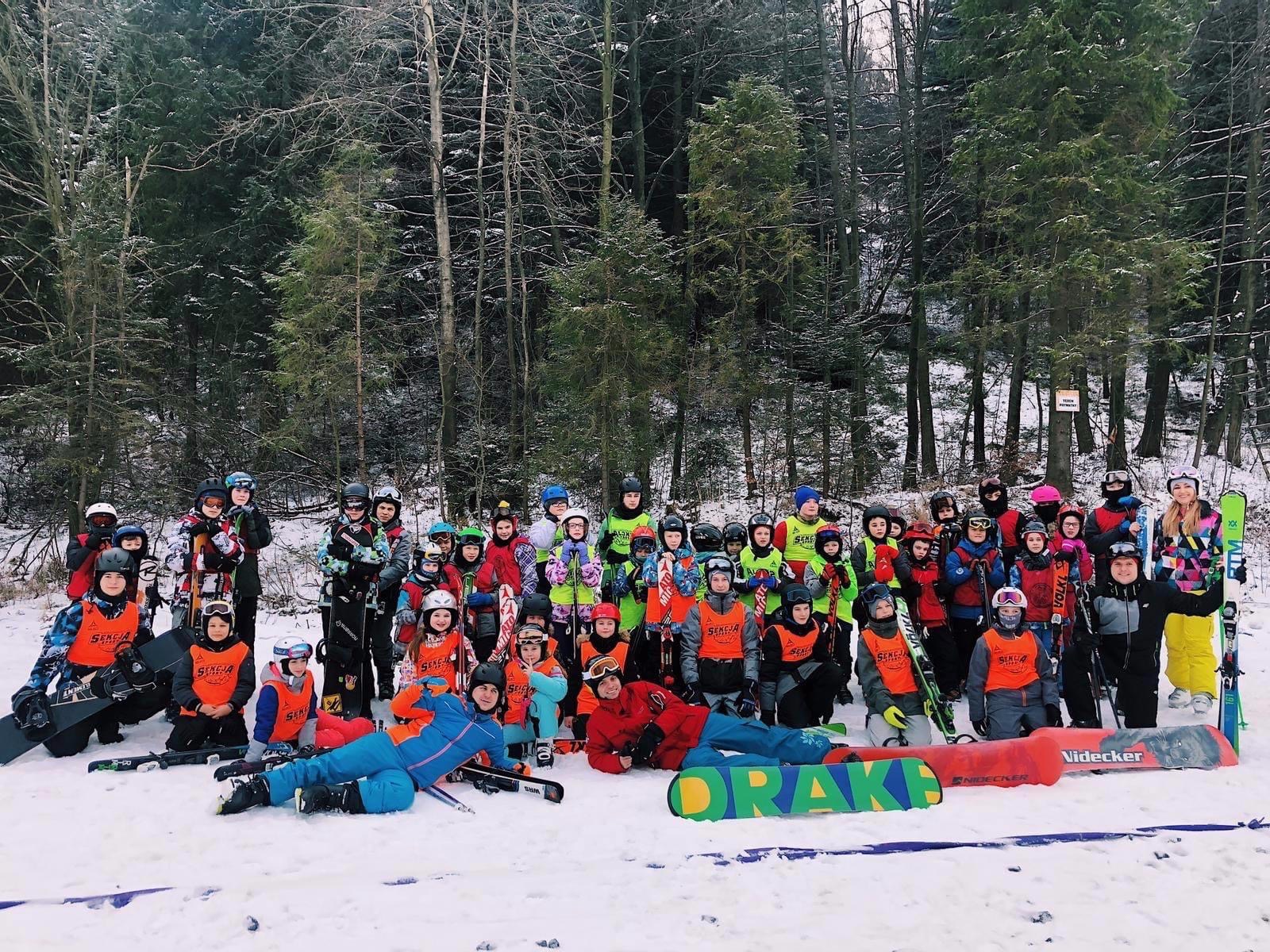 Duża grupa dzieci i dorosłych w kombinezonach narciarskich i kaskach pozuje do zdjęcia na tle lasu, trzymając narty i deski snowboardowe, na śniegu leżą deski z napisem DRAKE i NIDECKER.