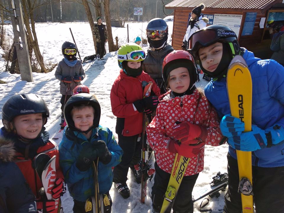 Grupa dzieci w kaskach i goglach narciarskich, trzymających narty, pozuje na tle zaśnieżonego stoku i budynku stacji narciarskiej.