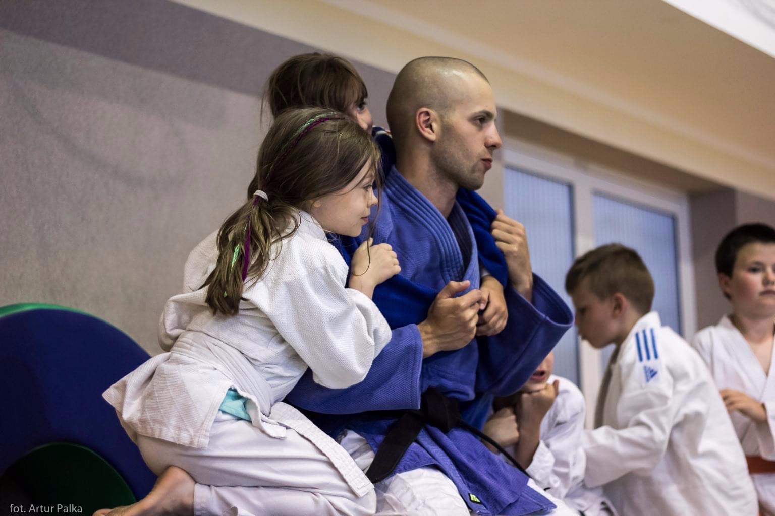 Instruktor judo w niebieskim stroju otoczony przez dzieci w białych kimonach, dziewczynka z kolorowymi pasemkami we włosach siedzi na jego kolanach.