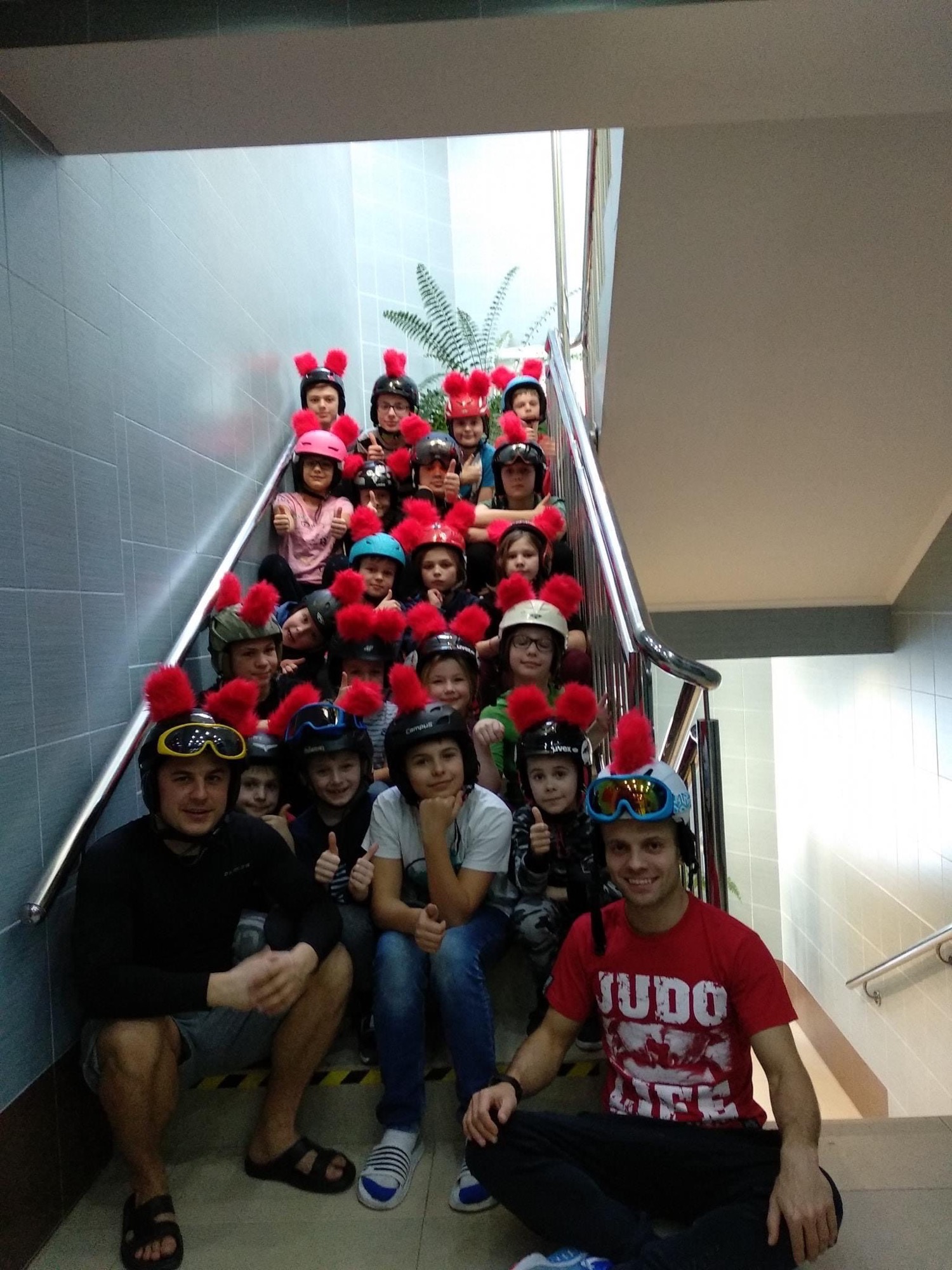 Grupa dzieci i dwóch mężczyzn w kaskach z czerwonymi pomponami siedzi na schodach; jeden z mężczyzn ma koszulkę z napisem 'JUDO LIFE', a dzieci pokazują kciuki w górę.